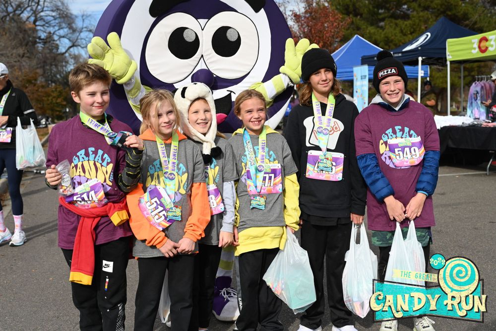 Five young runners pose with bags of candy in front of a giant lollipop-cosumed character.